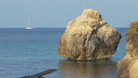 egg shaped rock in the mediterranean sea with a sailboat cruising on the water in the background