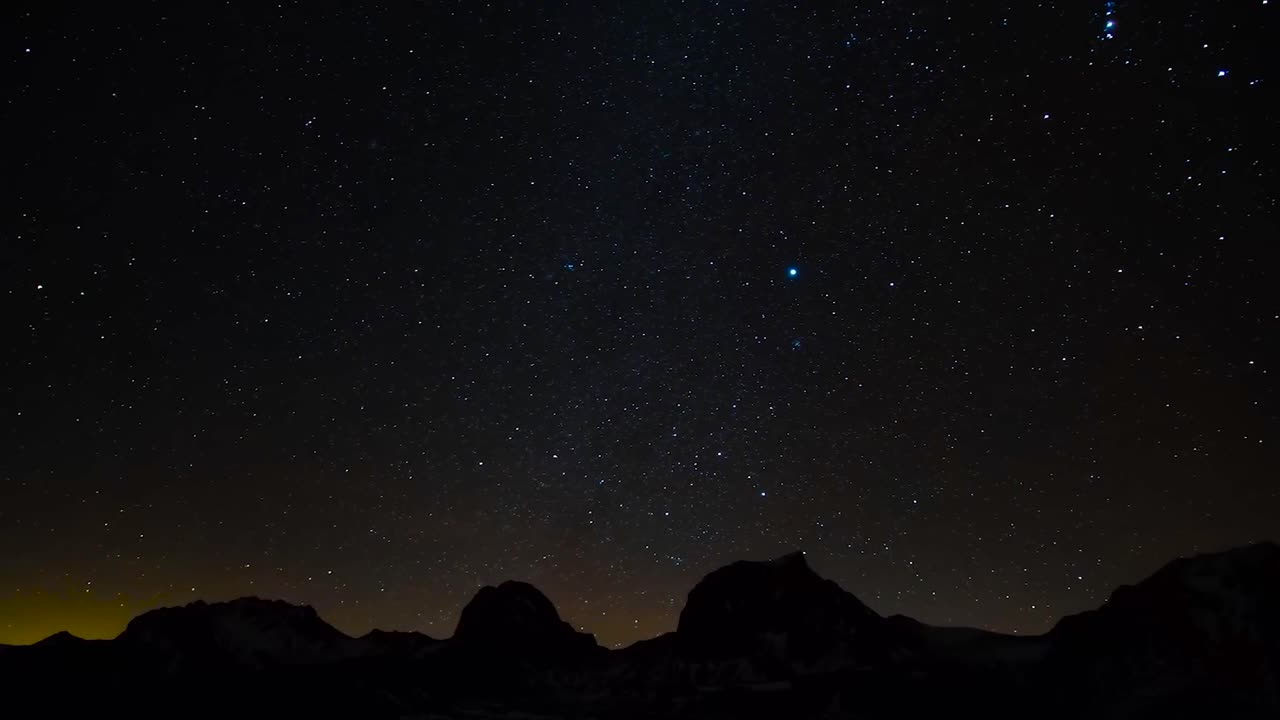 Time lapse or timelapse footage of a starry night or evening sky with bright stars. Mountain silhouettes are visible in the background and a small orange and yellow glare behind the mountains in 4K.