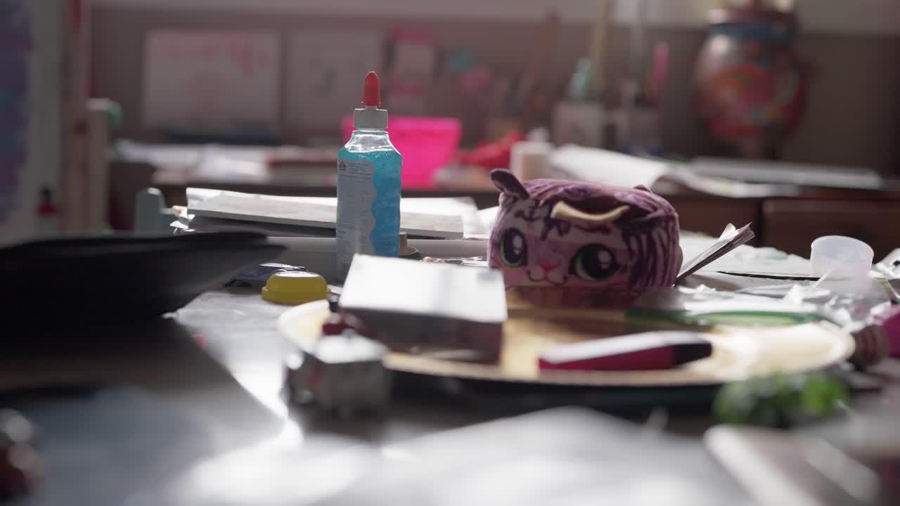 A stuffed cat animal is on a table with glue, papers and various arts and craft items.