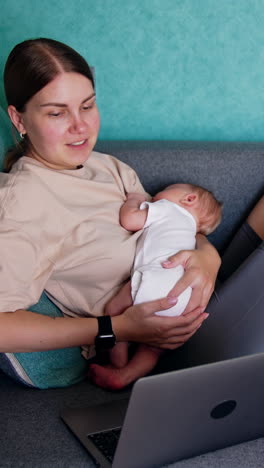 caucasian mother combining work and childcare. woman with child in arms works at laptop. vertical video
