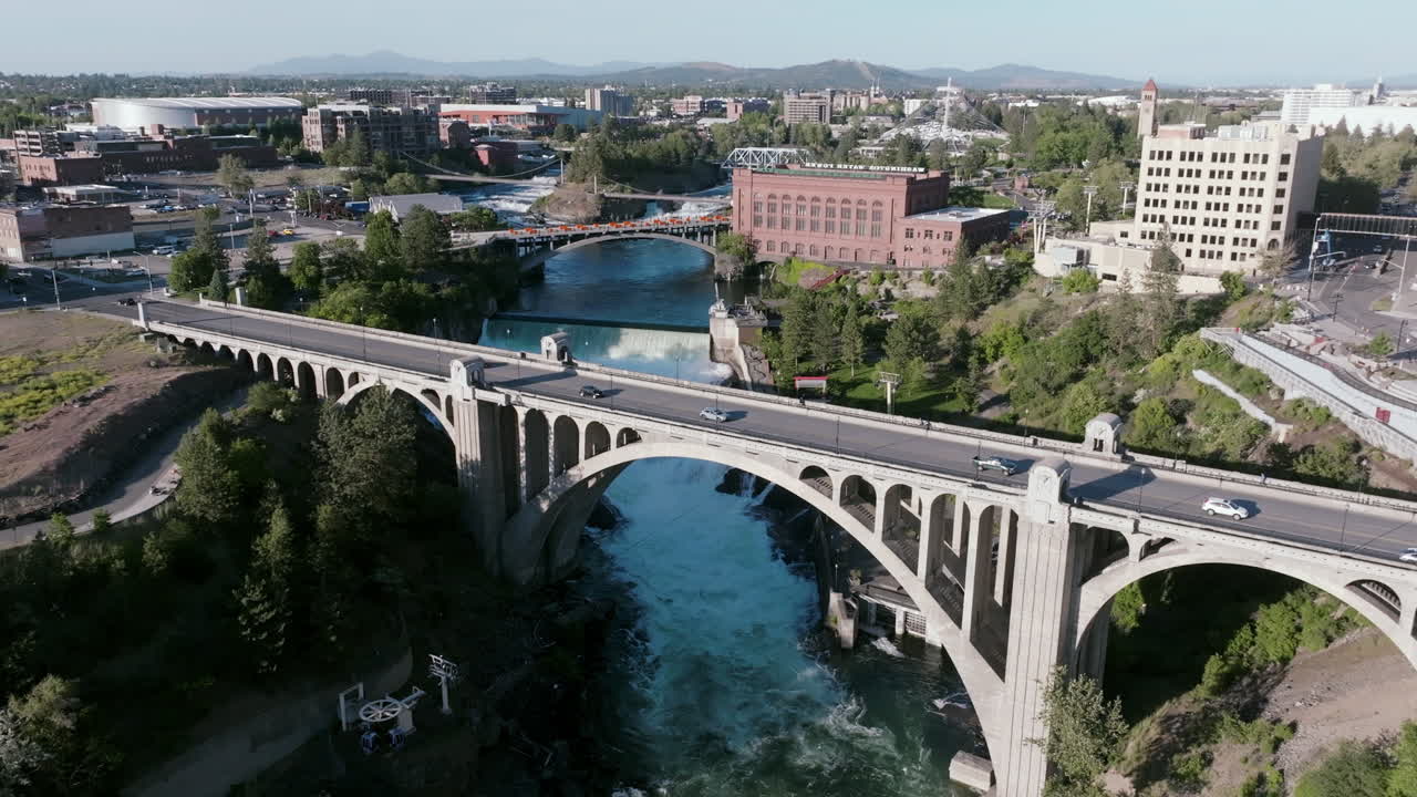 A wide shot of the Monroe Street Bridge arching gracefully over Spokane River's powerful flow, with city buildings and the cascading falls behind