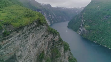 Aerial-Slomo-of-Geiranger-Fjord,-Norway,-flying-towards-a-Mountain-with-a-Moving-Boat-in-the-Background,-passing-The-Seven-Sisters