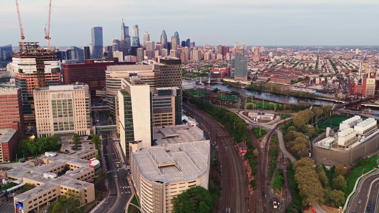 Aerial drone view straight down on multiple rail lines running between the Schuylkill River and a green cemetery with a commuter train visible on the tracks.