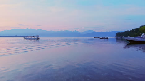 magical morning sunrise aerial of camera moving above calm reflective waters and past fishing boats in bali, indonesia with blue mountains and clear skies in the background