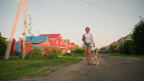 woman walking with her dog on leash along rural road, background featuring colorful buildings, greenery, and puddles on gravel path, displaying relaxed outdoor scene