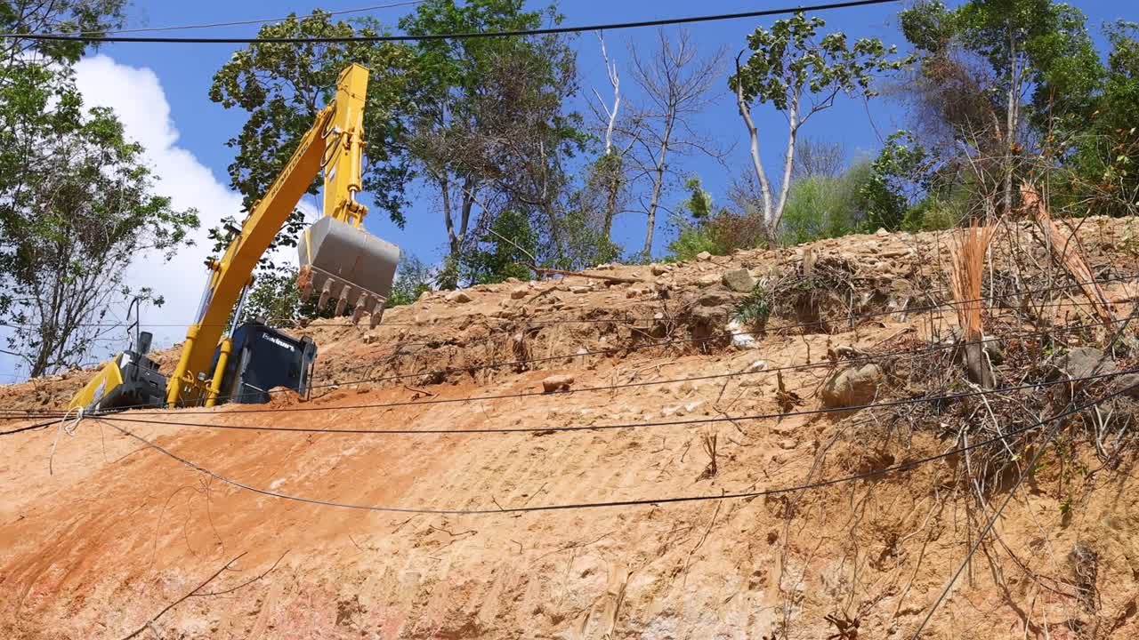 Excavator operates on a hillside under clear skies, moving earth with precision. Bright daylight enhances the vibrant natural surroundings