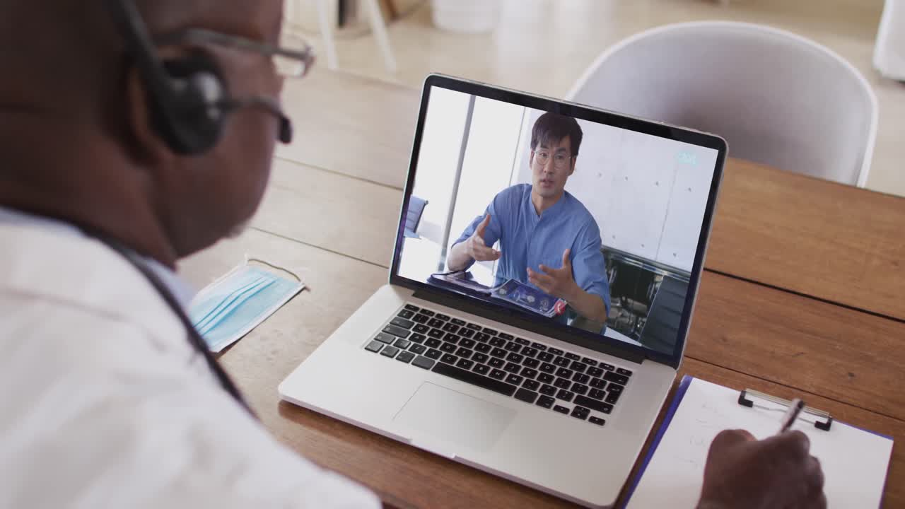 Ein afroamerikanischer Arzt mit Telefon-Headset macht sich Notizen während eines Videoanrufs am Laptop.