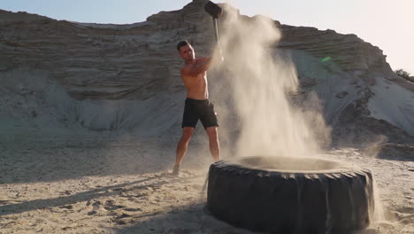 muscle athlete strongman man hits a hammer on a huge wheel in the sandy mountains in slow motion at sunset. the dust from the wheels rises.