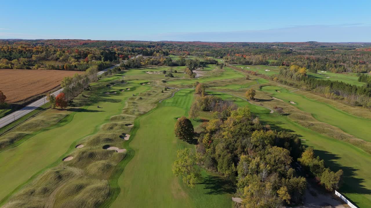 An expansive aerial view of a golf course surrounded by vibrant fall foliage. The landscape features rolling greens and a clear blue sky. Subtle camera roll movement