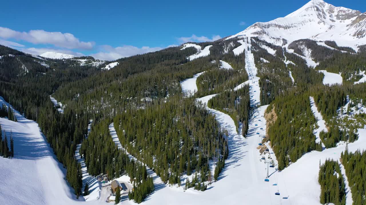 Drone flies over snowy Big Sky, Montana, showcasing a ski resort with skiers on chair lifts, carving down alpine trails surrounded by tall pine trees and dramatic winter mountain views