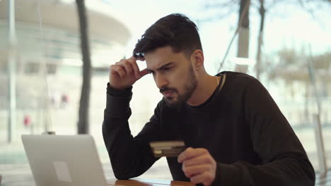 doubtful young man holding credit card and using laptop