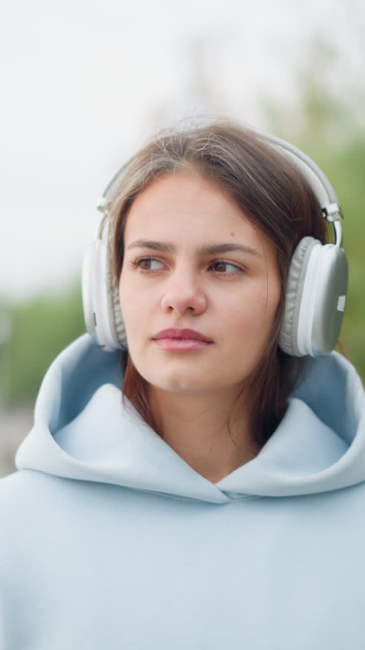 Close-up view of young woman strolling in garden wearing headphones, with blurred trees and bench in background. Perfect for videos about music enjoyment, relaxation, or nature walks