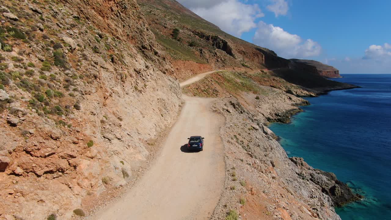 Crete Greece a dark blue car advances to Balos Beach on a seaside road to the remote location, Aerial follow shot with blurred plates