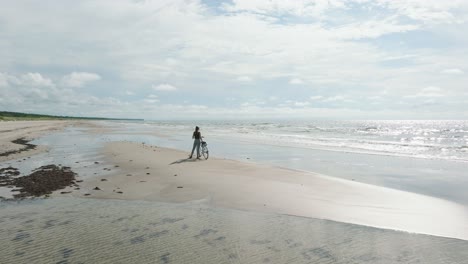 Aerial-view-with-a-young-longhaired-girl-standing-with-a-bike-on-the-sandy-beach,-sunny-day,-white-sand-beach,-active-lifestyle-concept,-low-wide-drone-shot-moving-forward