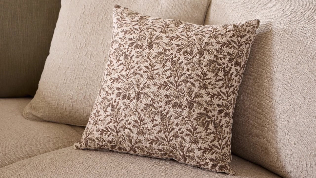 Lento zoom su un cuscino decorativo floreale appoggiato su un divano in lino beige-grigio, illuminato dalla calda luce del sole proveniente da una finestra fuori campo