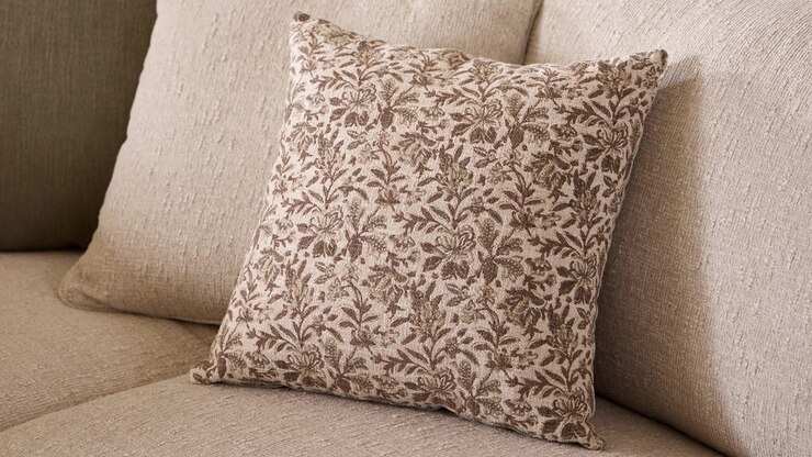 Lento zoom su un cuscino decorativo floreale appoggiato su un divano in lino beige-grigio, illuminato dalla calda luce del sole proveniente da una finestra fuori campo