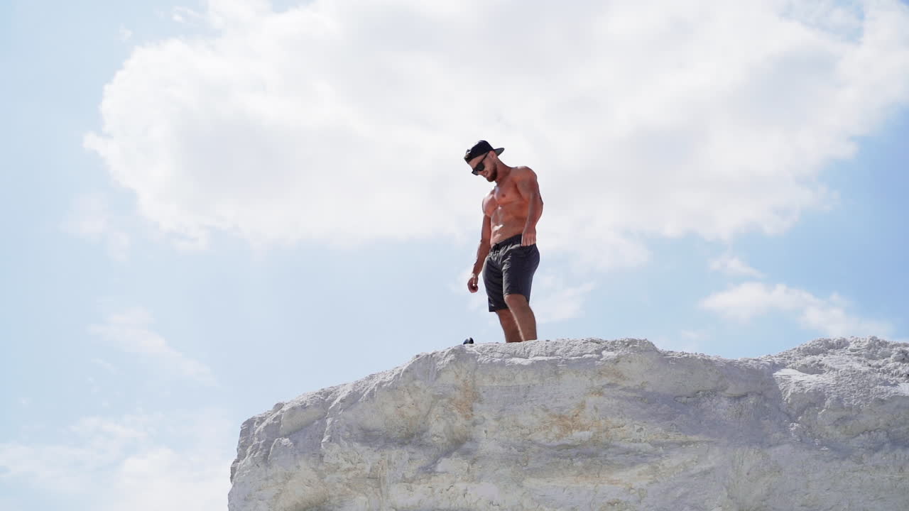 Shirtless athlete in cap and glasses on mountain. View from below on a sportsman standing on the top of white hill.