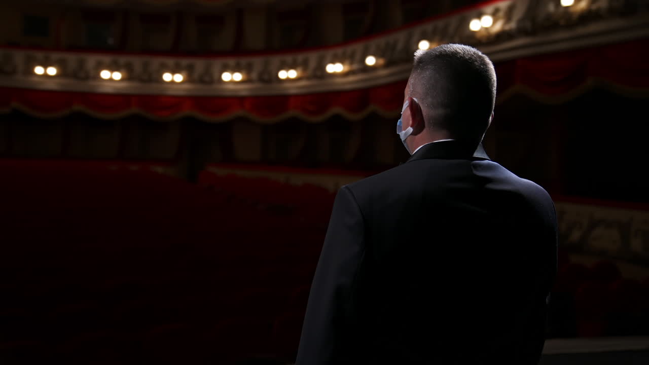 Performer man in front of empty auditorium. Man in suit taking off medical mask from is face and starts speaking while standing on stage in theater. Circling view.