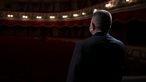 performer man in front of empty auditorium. man in suit taking off medical mask from is face and starts speaking while standing on stage in theater. circling view.