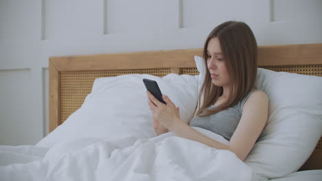 face close-up on woman typing a message on a mobile phone using 2 hands while comfortably lying on a bed in a hotel room. woman using smart phone and lying on the bed in morning after waking up