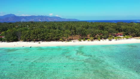 Quiet-beach-on-early-morning,-calm-clear-water-of-turquoise-lagoon-near-shore-of-tropical-island-with-lush-vegetation,-blue-sea-and-mountains-background