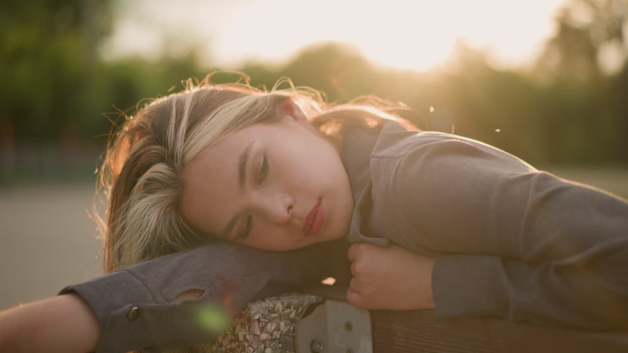 회색 옷을 입은 여자가 야외에서 팔에 머리를 고, 천천히 손을 내려, 조용한 표정으로 생각에 빠져, 흐릿한 배경은 부드러운 빛이 그녀를 둘러싸고있는 반사 상태를 강조합니다.