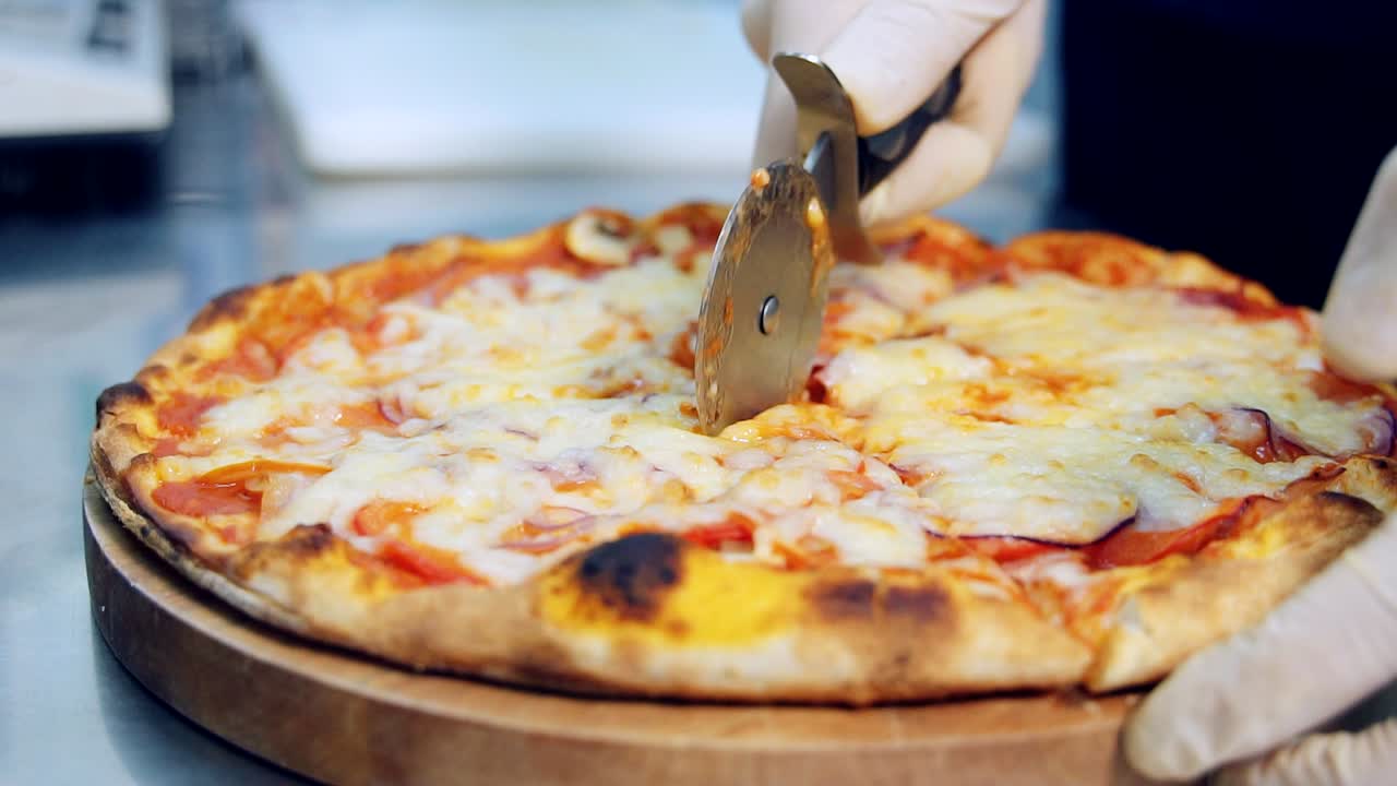 Pizza pie slicing into pieces. Cook using round cutter knife for cutting pizza on slices on wooden table close up