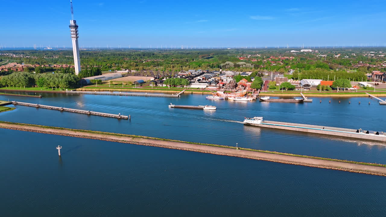 Barges move by the lake along the dam. Scenery of Lelystad, the Netherlands at backdrop. View on telecom tower and Batavialand museum.