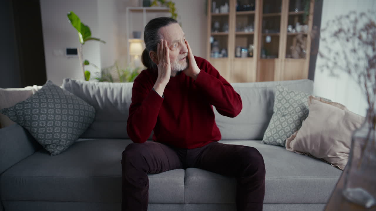 Elderly Man Sitting on Couch with Hands on Head Looking Distressed