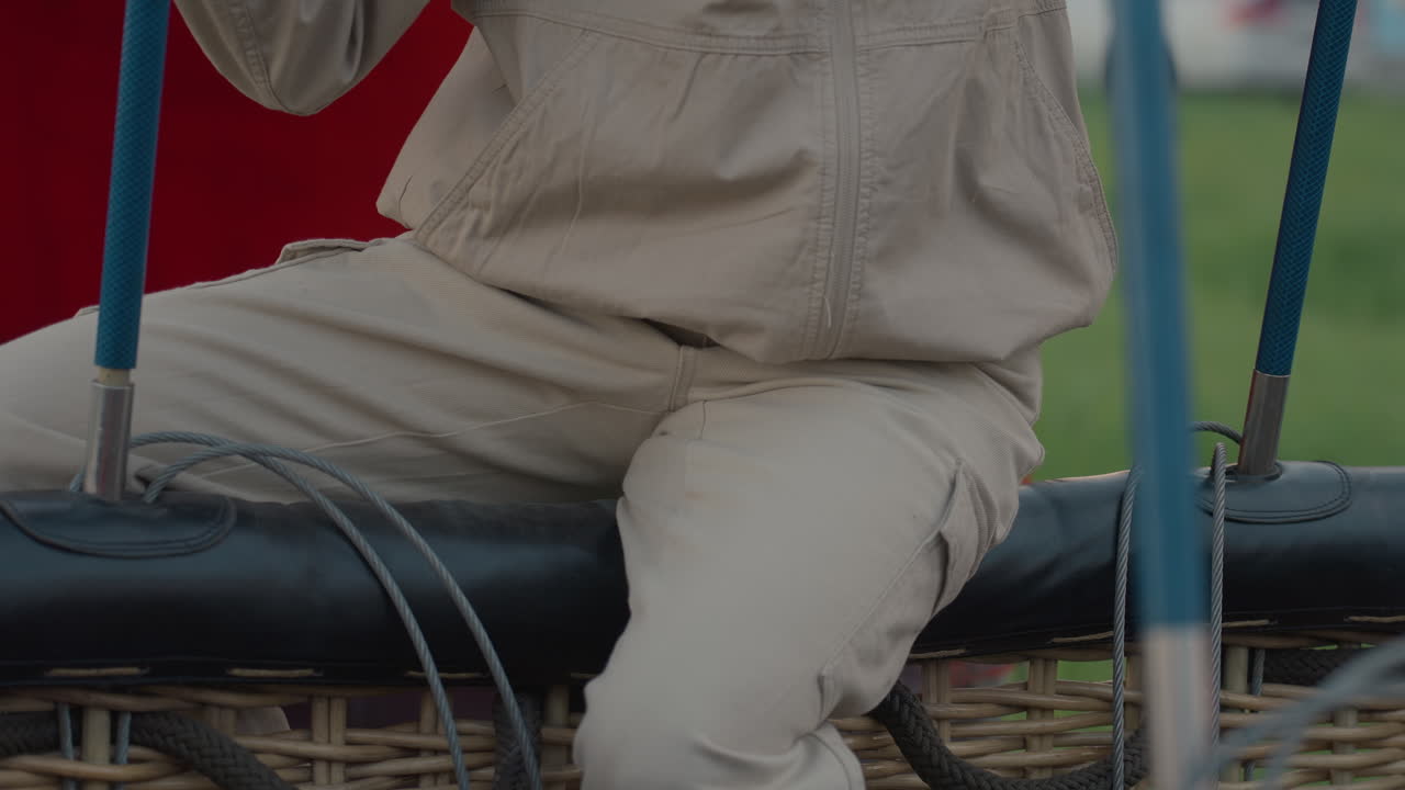 close up of person seated on wicker basket edge next to trailer rope and equipment before hot air balloon inflation in grassy field with parked car blurred background
