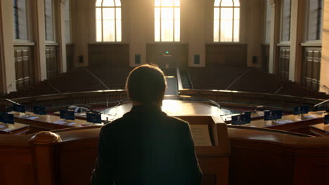 a person stands at a podium in an empty, grand assembly hall with strong backlighting