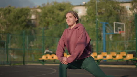 close-up of lady stretching in green leggings, pink hoodie, outdoor court background with sports equipment and seating area and trees with greenery