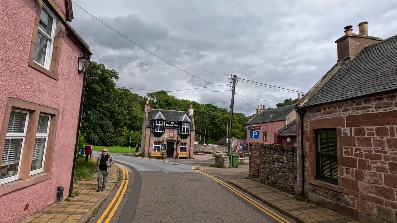 A person walks down a quiet, curved street lined with traditional pink stone houses in Cromarty, Scotland, under overcast daylight with steady camera movement
