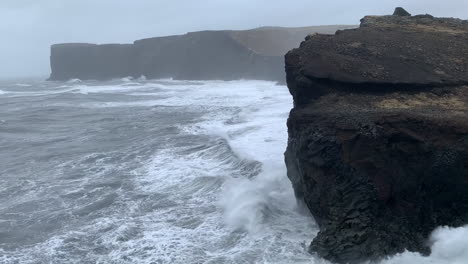 waves crash against jagged black cliffs on a misty coastline, sending sprays of white foam into the air. the overcast sky and rugged terrain highlight the dramatic seascape in iceland