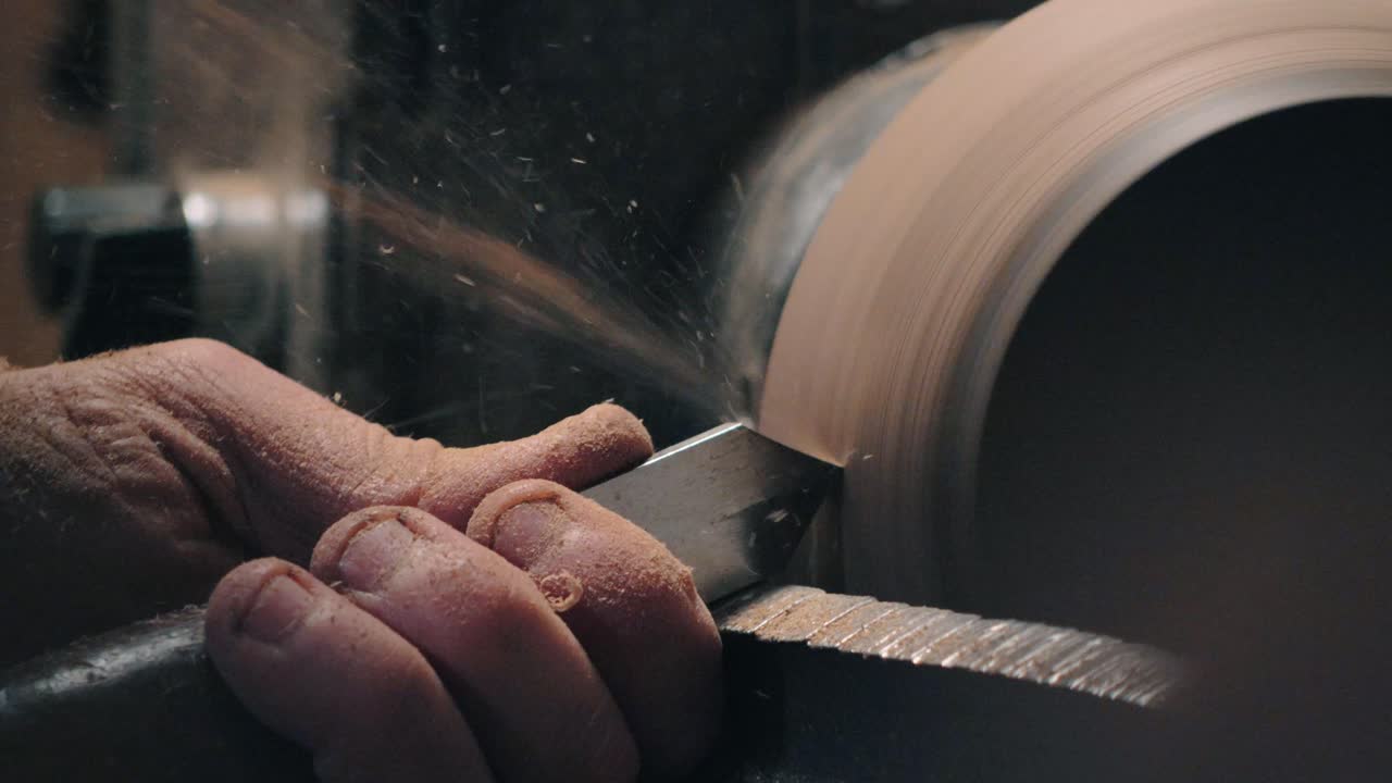 Woodturner hands guiding a chisel against spinning wood on a lathe. Flying shavings and sawdust highlight the woodturning process in an artisan workshop
