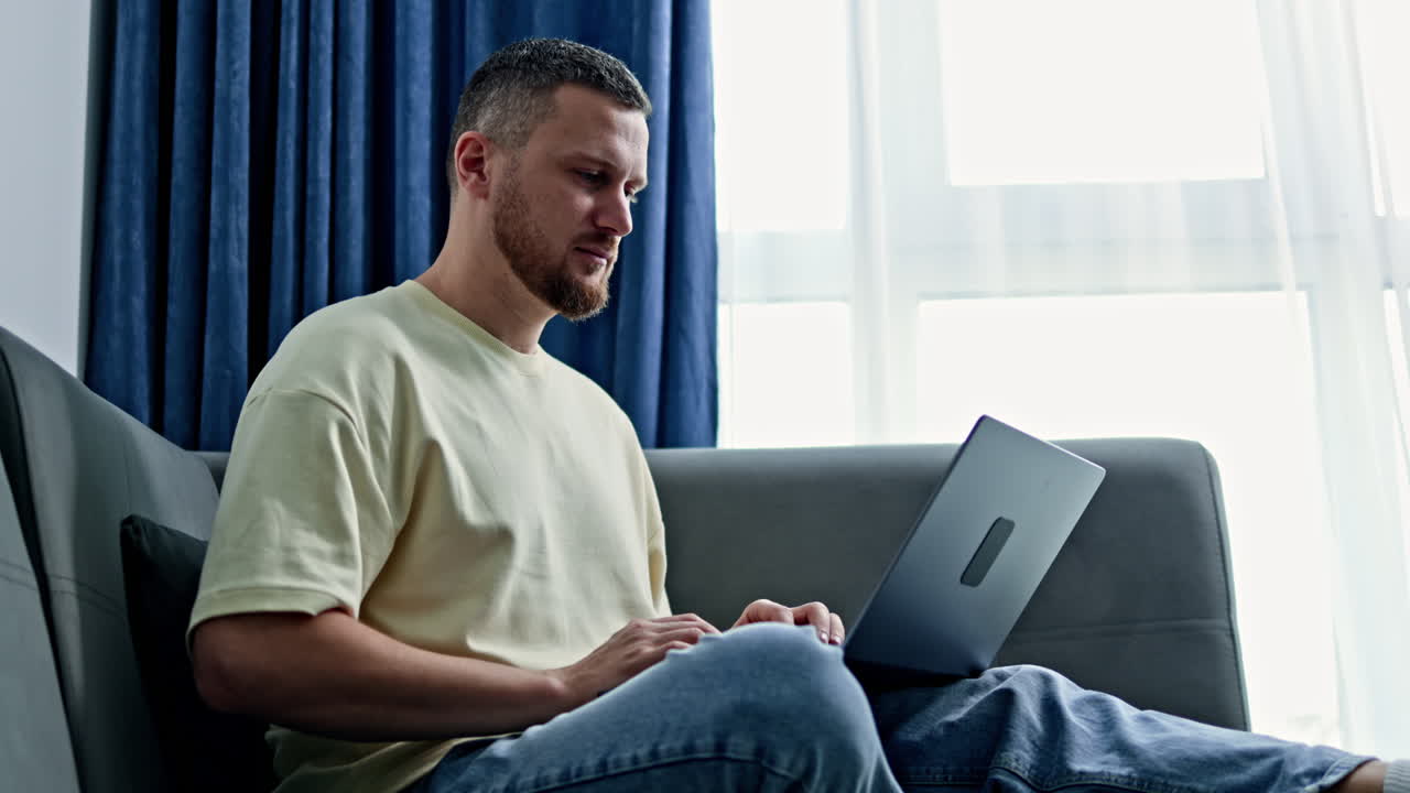Person works on laptop while sitting on sofa near window in modern room during day time