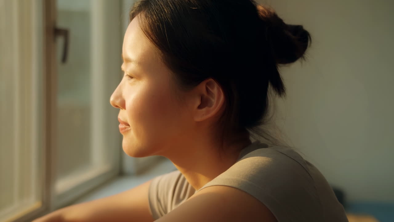 朝の光が差し込む窓辺の女性。顔に当たる優しい日差しと物思いにふける表情、静かなアパートの一室の情景。