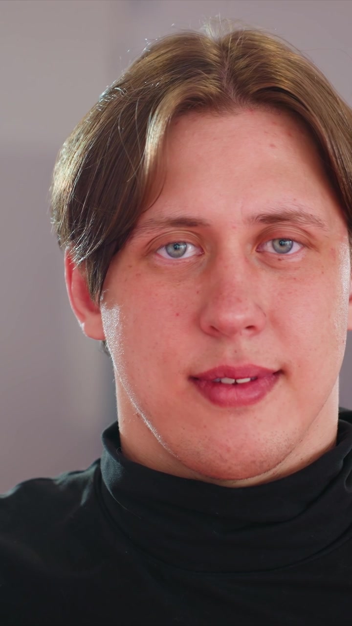 Primo piano di un giovane uomo con capelli castano chiaro. La sua testa si muove leggermente, rivelando brevemente l'orecchio di un altro uomo. Lo sfondo è semplice e neutro per tutta la durata, mantenendo un aspetto uniforme.