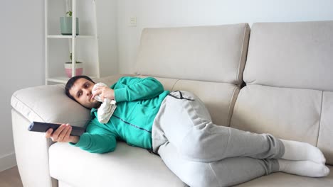 hombre con dolor de dientes acostado en el sofá mientras enciende la tv y sostiene la mejilla en el dolor