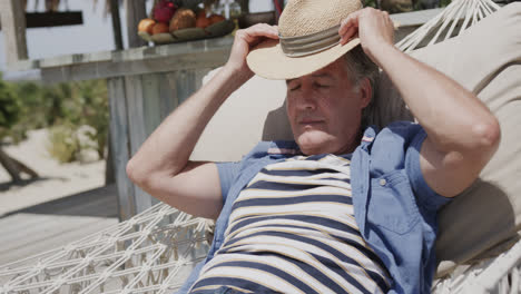 happy senior caucasian man in hat relaxing in hammock in the sun by beach bar, in slow motion