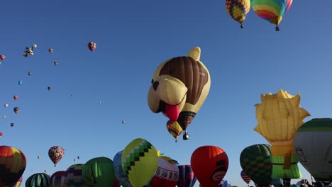 Happy-puppy-head-hot-air-balloon-floating-over-a-row-of-colorful-balloons-with-ascending-balloons-in-the-background-on-a-clear-early-morning-mass-ascension-in-slow-motion