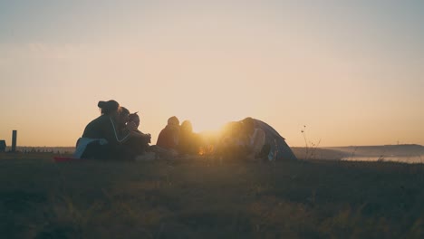 young people rest at burning bonfire on river bank at sunset