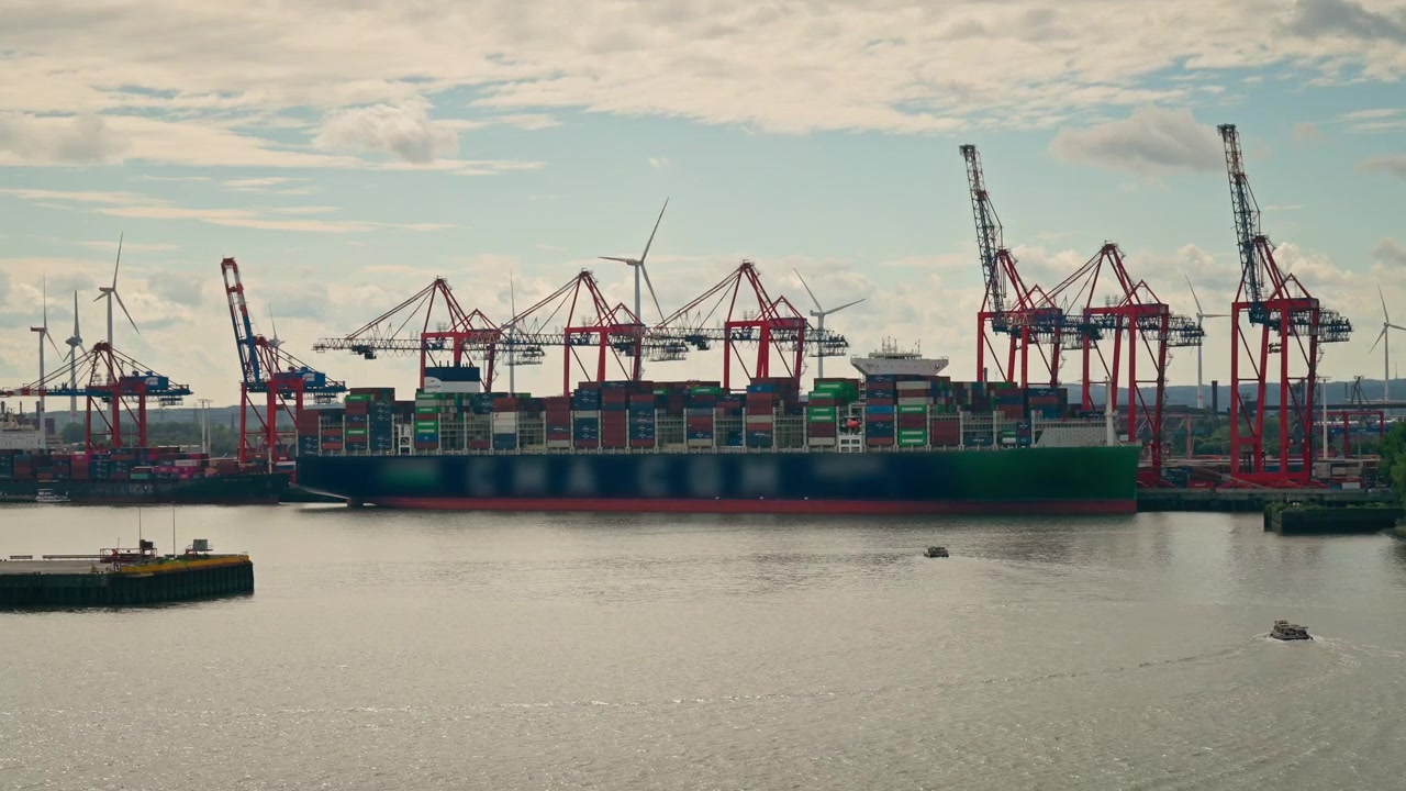 Large cargo ship moored beside rows of container gantry cranes and stacked freight containers, with wind turbines in the background at a busy industrial waterfront.