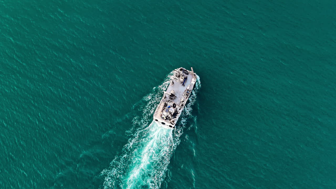 Piccola imbarcazione industriale che si muove attraverso acqua marina limpida e turchese creando una scia di schiuma bianca. Vista aerea dall'alto di una barca da pesca in acqua turchese.
