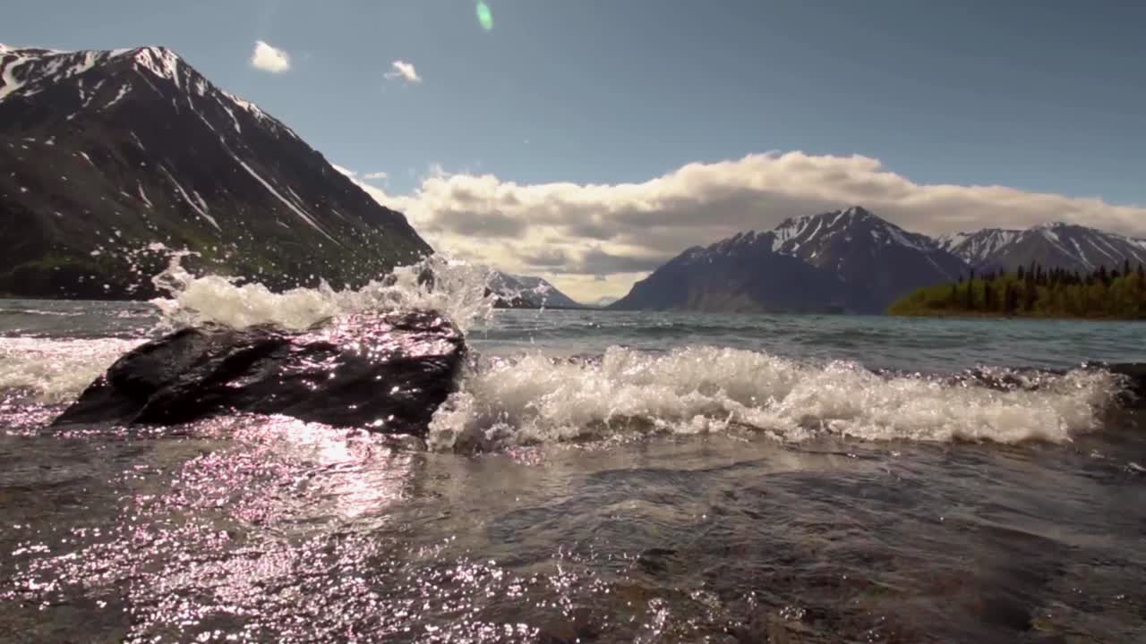 Low wide angle view of Yukon Kathleen lake waves crashing and flowing towards camera and past rock in water by scenic mountain countryside range on sunny day and sun flare, Canada, close up handheld