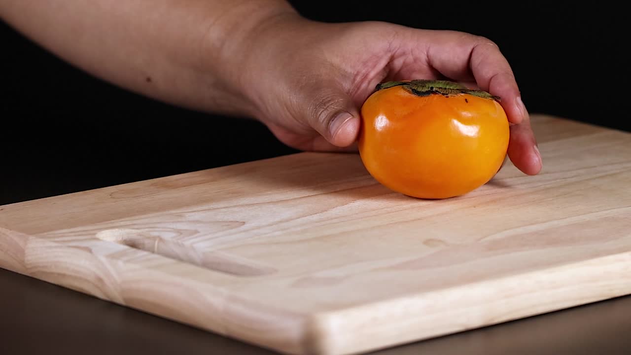 A hand carefully places an orange persimmon on a light wooden cutting board against a dark background.