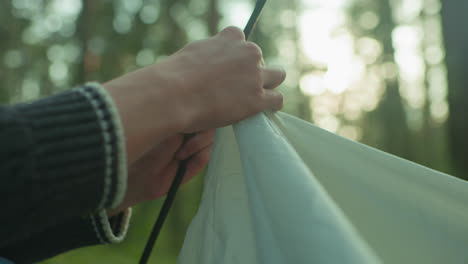 close up of person wearing knitted sweater carefully holding tent pole and tent fabric while assembling outdoor shelter in forest with soft daylight creating warm natural background