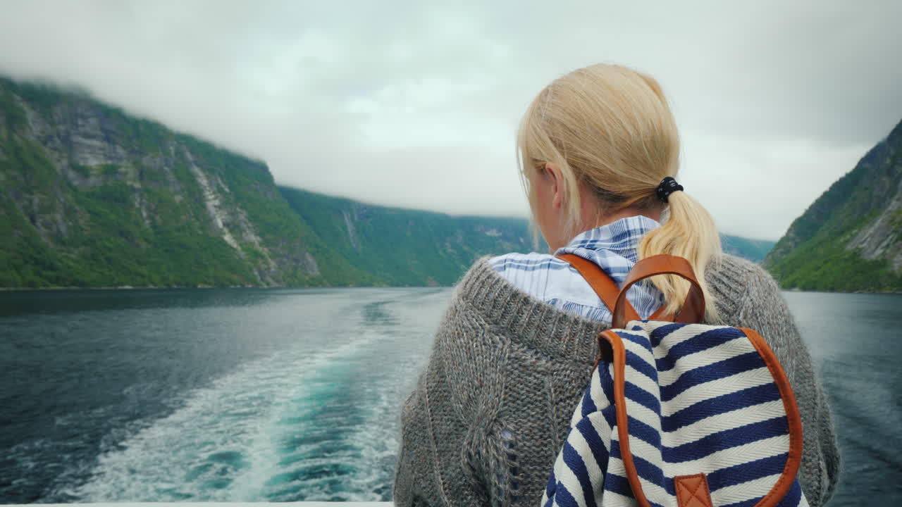 한 여성이 유람선 선미에 서서 후퇴하는 바위와 피요르드의 파도를 바라보고 있습니다.