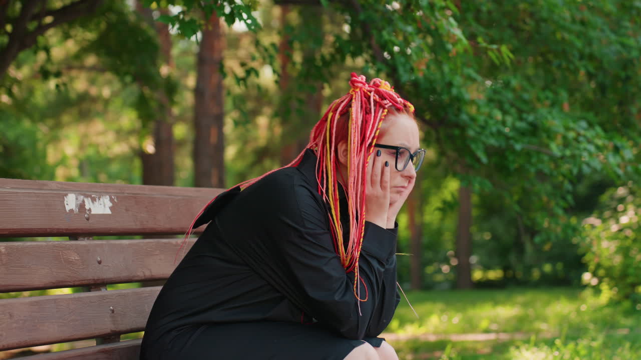 Белая женщина с пастельными косами стоит и сидит возле парковой скамейки с закрытыми глазами и спокойным выражением лица, тихо дышит, сосредоточившись на себе, листва на заднем плане, тихая стойкость