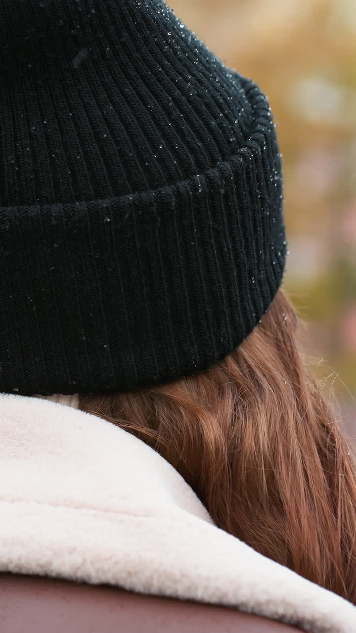 Close back view of young girl wearing black knit cap and brown shearling jacket walking outdoors during overcast day, blurred urban park background adds calm peaceful atmosphere